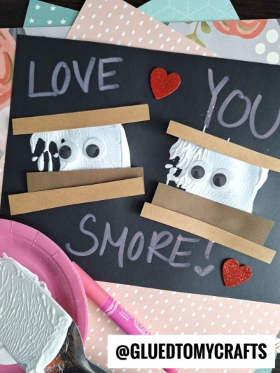 A spatula stamped smores craft card with LOVE YOU SMORE! written in chalk on black paper, featuring two smores made from brown strips, white paint, googly eyes, and red glitter hearts. Craft supplies are scattered around the card.