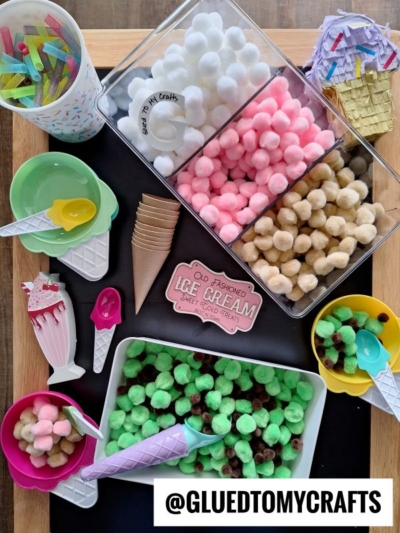 Colorful craft supplies arranged to look like ice cream, including pom-poms in white, pink, tan, and green, mini cones, colorful spoons, bowls, straws, and a sign reading “Go Fashioned Ice Cream.”.