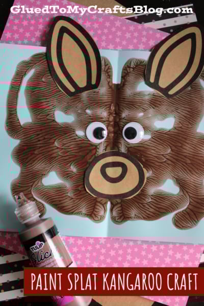 A paint splat craft resembling a kangaroo with googly eyes, large paper ears, and a round nose on blue paper. A bottle of brown fabric paint and polka dot paper are nearby. Text reads “Paint Splat Kangaroo Craft.”.