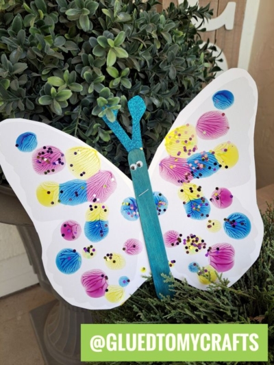 A colorful butterfly PAINT SPLAT CRAFT made from a white paper cutout, decorated with painted pasta shells and sequins, with a blue craft stick body and googly eyes, displayed among green plants. @GLUEDTOMYCRAFTS is written at the bottom.