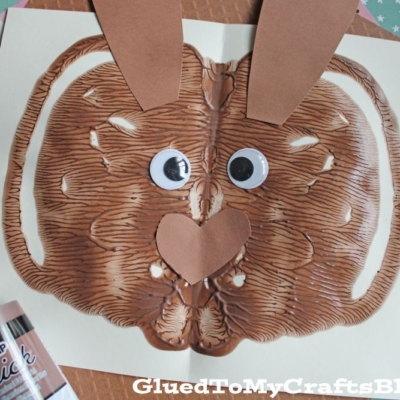 A brown paint blot folded on white paper to resemble a bunny face, with googly eyes, a paper nose and ears, and brown puffy paint nearby. The background shows textured and patterned paper.