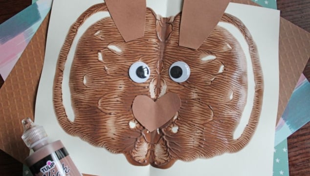 A brown paint blot folded on white paper to resemble a bunny face, with googly eyes, a paper nose and ears, and brown puffy paint nearby. The background shows textured and patterned paper.