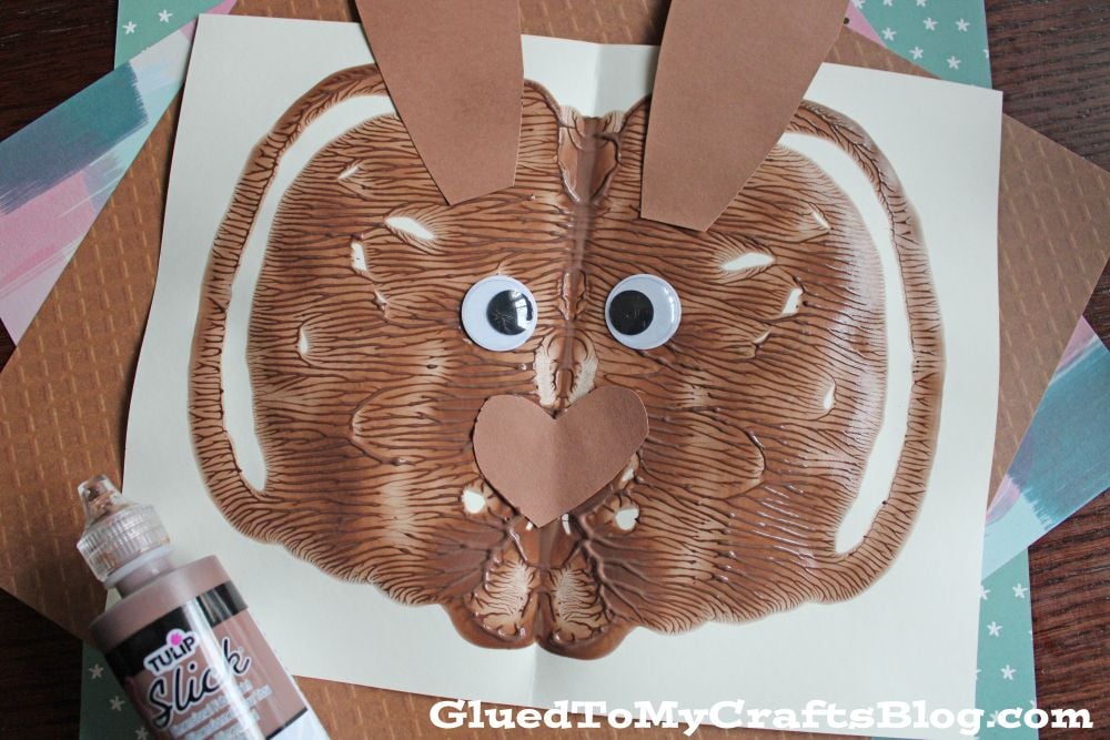 A brown paint blot folded on white paper to resemble a bunny face, with googly eyes, a paper nose and ears, and brown puffy paint nearby. The background shows textured and patterned paper.