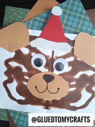 A festive PAINT SPLAT CRAFTS Christmas bear made with brown paint, paper cutouts for ears, nose, and mouth, large googly eyes, and a red Santa hat, displayed on green and brown patterned paper. @GLUEDTOMYCRAFTS is written at the bottom.
