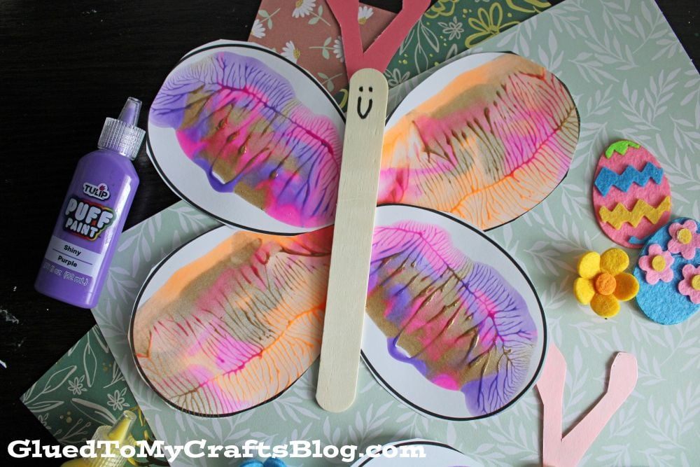 A colorful butterfly craft made from painted paper wings, a wooden stick for the body with a smiley face, and decorated with flowers and Easter egg cutouts. A bottle of purple puff paint is nearby.