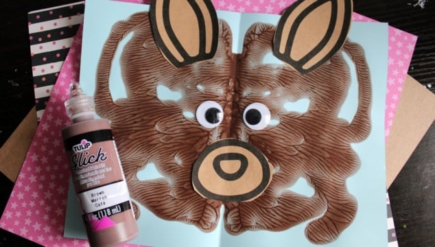 A brown painted kangaroo face craft with large googly eyes and paper ears is displayed on blue paper, next to a bottle of brown fabric paint. The background includes pink and black patterned papers.