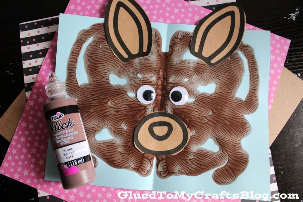 A brown painted kangaroo face craft with large googly eyes and paper ears is displayed on blue paper, next to a bottle of brown fabric paint. The background includes pink and black patterned papers.