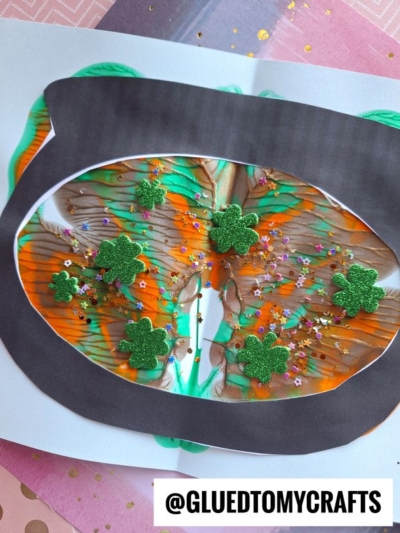 A colorful St. Patrick’s Day craft features a paper pot of gold with orange, green, and brown PAINT SPLAT CRAFTS effects, glitter, and green shamrock decorations. The username @GLUEDTOMYCRAFTS appears at the bottom.