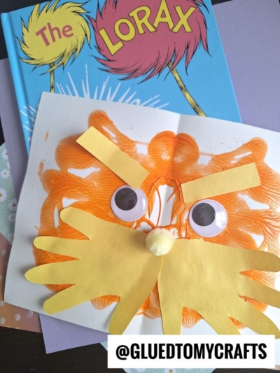 A “The Lorax” book with a homemade Lorax face craft on top, featuring orange paint, googly eyes, and yellow paper cut-outs for eyebrows and mustache, plus a yellow pom-pom for the nose. @GLUEDTOMYCRAFTS is labeled at the bottom.