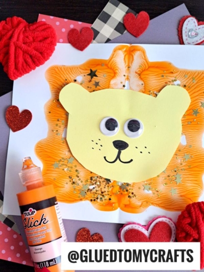 A handmade lion paint splat craft features a yellow paper face, large googly eyes, and orange paint spread behind it. Surrounded by craft supplies, glitter, and heart decorations. The username @GLUEDTOMYCRAFTS is visible.