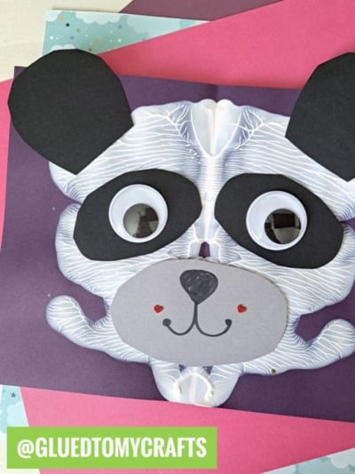 A paint splat panda bear face craft made with black and gray paper shapes, large googly eyes, and drawn facial features, is displayed on colorful sheets. The handle @GLUEDTOMYCRAFTS is written in green at the bottom.
