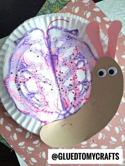 A paper plate PAINT SPLAT CRAFT decorated with purple and pink paint, glitter, and a construction paper snail face with googly eyes and pink antennae, placed on floral-patterned paper.