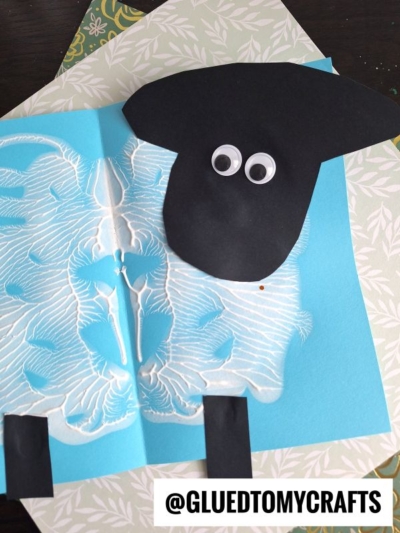 A PAINT SPLAT CRAFTS paper sheep made with blue and white paint, black paper for the head and legs, and two googly eyes, displayed on a table with patterned paper. The handle @GLUEDTOMYCRAFTS is shown.