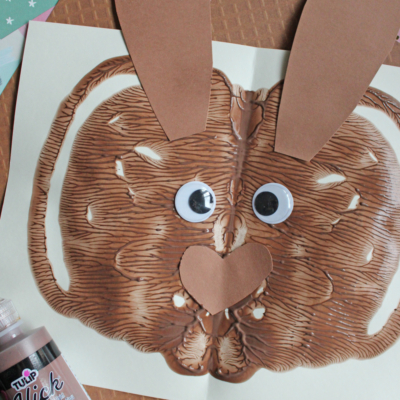A paint splat creating a chocolate-colored bunny face on paper, with added large googly eyes, brown paper ears, and a heart-shaped nose. A bottle of fabric paint and papers are in the background. Text reads: Paint Splat Chocolate Bunny.