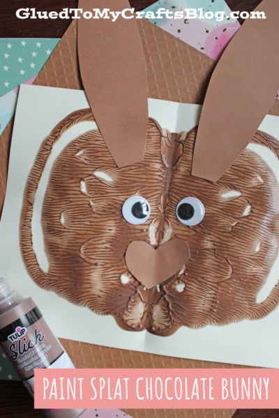 A paint splat creating a chocolate-colored bunny face on paper, with added large googly eyes, brown paper ears, and a heart-shaped nose. A bottle of fabric paint and papers are in the background. Text reads: Paint Splat Chocolate Bunny.