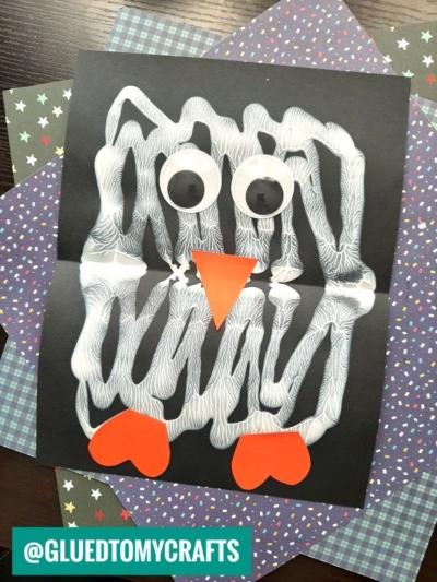 A kids’ PAINT SPLAT CRAFT of a penguin made with white painted zigzags on black paper, large googly eyes, an orange paper beak, and two orange paper feet, displayed on patterned paper.