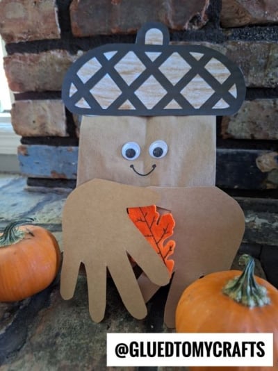 A paper bag craft shaped like an acorn with googly eyes and a smile, decorated with a handprint cutout and felt leaf, sits between two small pumpkins—one of the cutest PAPER BAG CRAFT IDEAS FOR FALL. The handle @GLUEDTOMYCRAFTS is visible at the bottom.