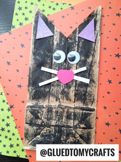A paper bag decorated as a cat with black coloring, googly eyes, white paper whiskers, a pink heart nose, and purple glitter ears—perfect PAPER BAG CRAFT IDEAS FOR FALL—placed on orange paper with black stars. Text reads @GLUEDTOMYCRAFTS.