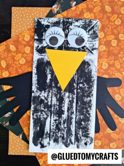 A paper bag crow craft with googly eyes, a yellow beak, and black paper wings on orange patterned paper. The handle @GLUEDTOMYCRAFTS appears in the bottom right corner.