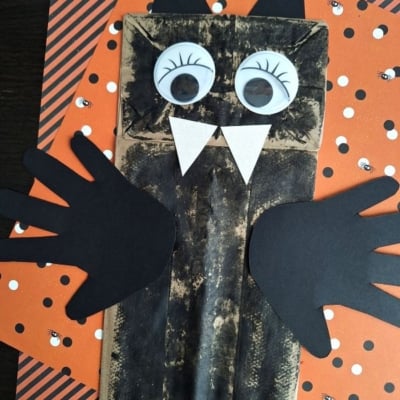 A fun PAPER BAG CRAFT IDEA FOR FALL, this bat craft is made from a brown paper bag with black wings and ears, big googly eyes, white fangs, and set on orange Halloween-themed paper.
