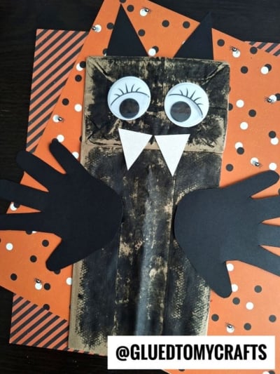A fun PAPER BAG CRAFT IDEA FOR FALL, this bat craft is made from a brown paper bag with black wings and ears, big googly eyes, white fangs, and set on orange Halloween-themed paper.
