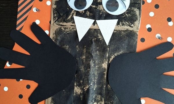 A fun PAPER BAG CRAFT IDEA FOR FALL, this bat craft is made from a brown paper bag with black wings and ears, big googly eyes, white fangs, and set on orange Halloween-themed paper.