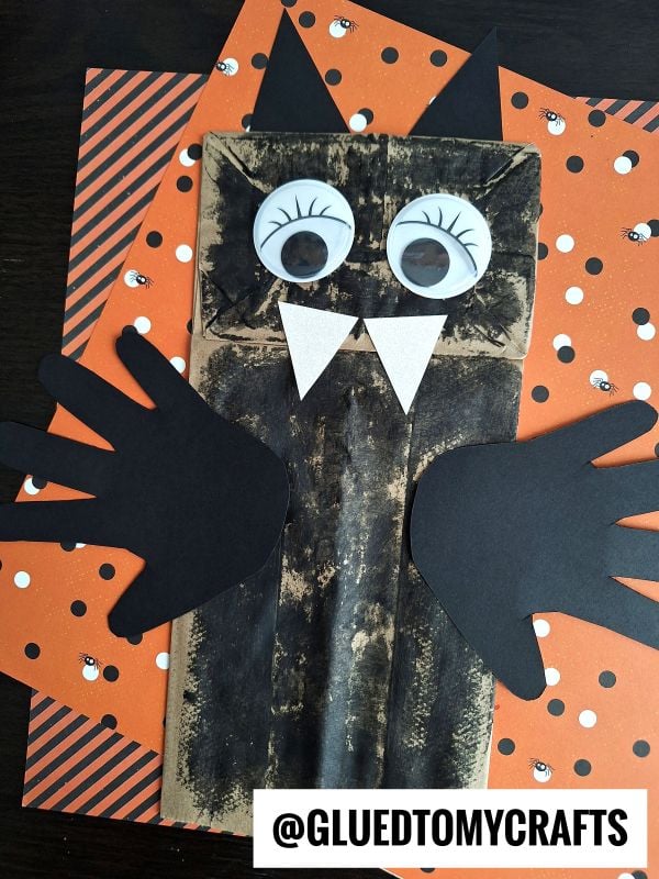 A fun PAPER BAG CRAFT IDEA FOR FALL, this bat craft is made from a brown paper bag with black wings and ears, big googly eyes, white fangs, and set on orange Halloween-themed paper.