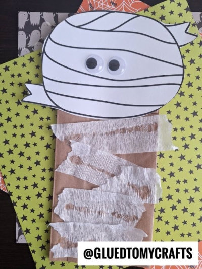 A paper bag decorated as a mummy with white strips and googly eyes on a patterned yellow and orange background. The text @GLUEDTOMYCRAFTS appears at the bottom right.