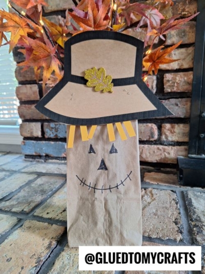 A paper bag scarecrow craft with a drawn face, yellow paper strips as hair, and a black-trimmed hat decorated with a glittery gold leaf, sits on a brick surface with autumn leaves in the background.