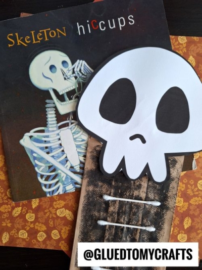 Flat lay of the book “Skeleton Hiccups,” a skeleton craft made from paper and cotton swabs, and an orange patterned background. The handle @GLUEDTOMYCRAFTS is at the bottom.