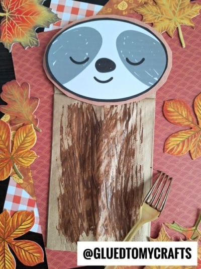 A sloth craft made from a painted brown paper bag with a smiling sloth face attached on top, surrounded by autumn leaves and a gold plastic fork. @GLUETOMYCRAFTS is written at the bottom.