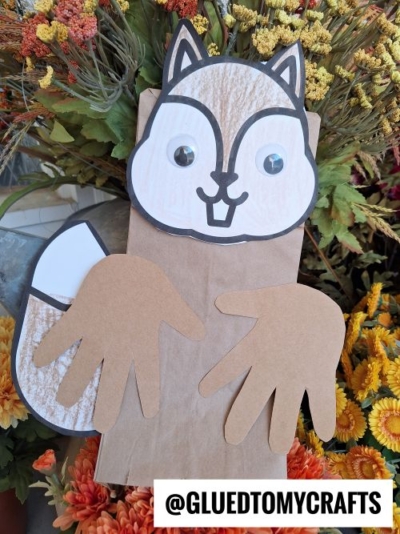 A squirrel puppet made from a brown paper bag, with a paper face, googly eyes, and hand-shaped cutouts for arms, is surrounded by colorful flowers. Text at the bottom reads @GLUEDTOMYCRAFTS.