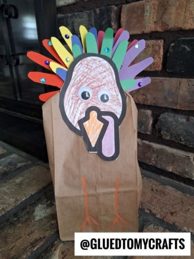 A brown paper bag decorated as a turkey with colorful paper feathers, googly eyes, and a drawn beak and wattle; orange legs are drawn on the bag. The handle @GLUEDTOMYCRAFTS is shown at the bottom.