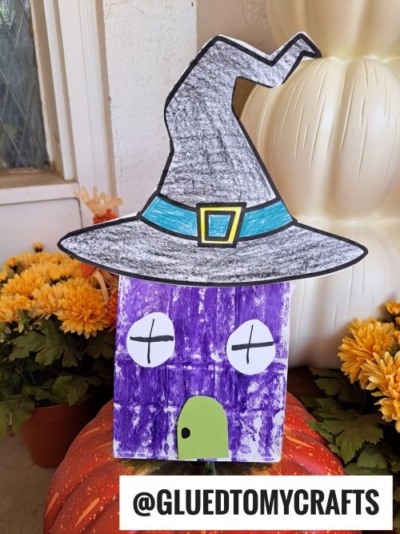 A purple paper bag decorated as a witch’s house with a large witch hat, round eyes, and a green door. The house sits among orange pumpkins and yellow flowers. @GLUEDTOMYCRAFTS is written at the bottom.