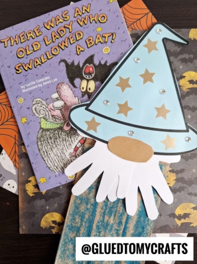 A children’s book titled “There Was an Old Lady Who Swallowed a Bat!” lies next to a handmade wizard craft with a blue hat and white beard, on a Halloween-themed background. The handle @gluedtomycrafts appears at the bottom.