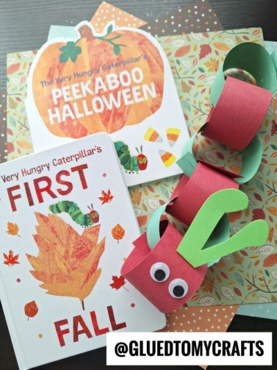 Two Very Hungry Caterpillar books, fall-themed scrapbook paper, and a HUNGRY CATERPILLAR CRAFT made from red and green paper rings with googly eyes are arranged together. A label reads “@GLUEDTOMYCRAFTS.”.