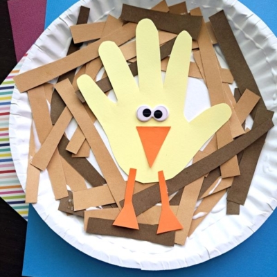 A paper plate craft featuring a yellow handprint turkey with googly eyes, an orange beak, and legs, surrounded by brown paper strips, on top of colored paper. Text at the bottom reads @GLUEDTOMYCRAFTS.