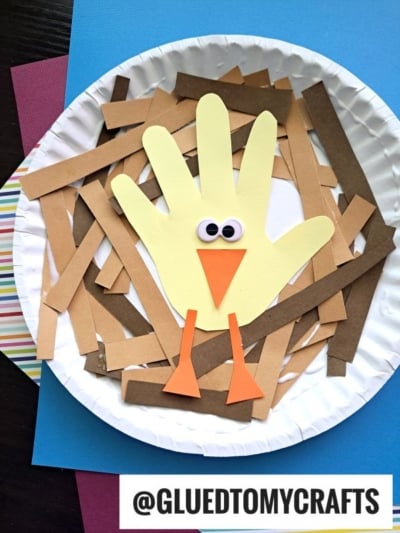 A paper plate craft featuring a yellow handprint turkey with googly eyes, an orange beak, and legs, surrounded by brown paper strips, on top of colored paper. Text at the bottom reads @GLUEDTOMYCRAFTS.