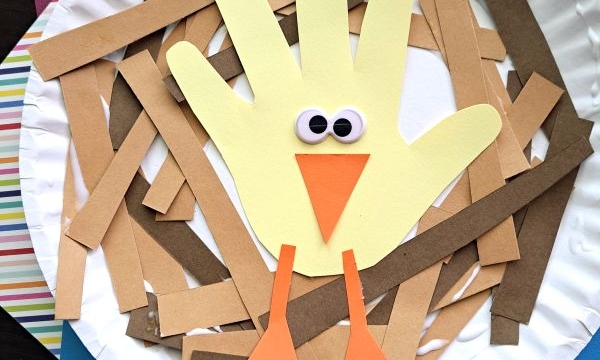 A paper plate craft featuring a yellow handprint turkey with googly eyes, an orange beak, and legs, surrounded by brown paper strips, on top of colored paper. Text at the bottom reads @GLUEDTOMYCRAFTS.