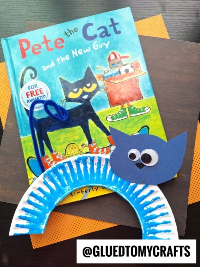 A copy of Pete the Cat and the New Guy sits on orange paper. In front is a blue cat craft made from a paper plate and construction paper—perfect for pete the cat crafts fans! The handle @GLUEDTOMYCRAFTS appears at the bottom.