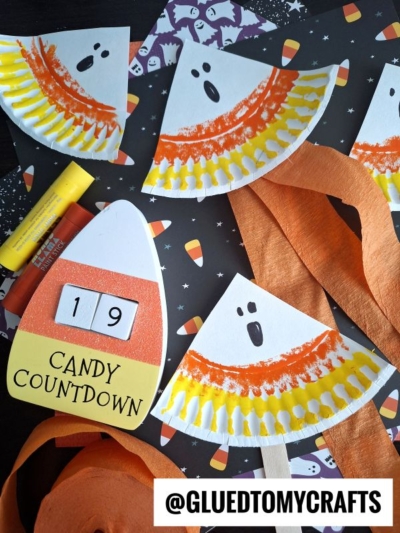 Paper plate candy corn ghosts decorated with orange and yellow colors are arranged on a Halloween-themed background featuring candy corn patterns, a candy countdown calendar showing “19,” and festive orange streamers.