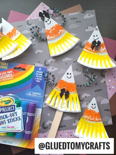Four paper plate candy corn crafts with smiling faces and pipe cleaner arms are arranged on star and moon-themed paper, next to a pack of Crayola paint sticks. A “@GLUEDTOMYCRAFTS” tag appears at the bottom.