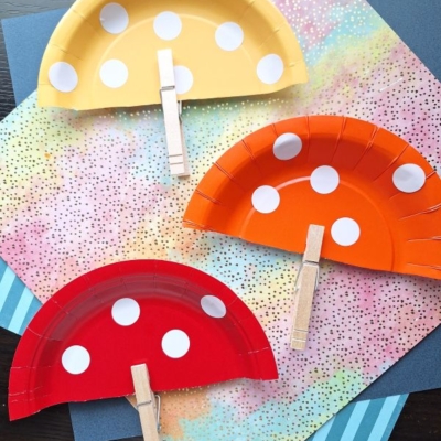 Three paper plate fans, each cut in half and painted yellow, orange, and red with white polka dots like a clothespin mushroom, have clothespins attached as handles. They’re displayed on colorful, patterned paper backgrounds.