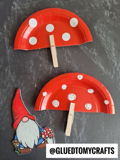 Two red paper plate halves decorated as mushrooms with white dots and clothespin stems are placed on a black surface next to a cutout drawing of a gnome holding a mushroom. Text at the bottom reads @GLUEDTOMYCRAFTS.