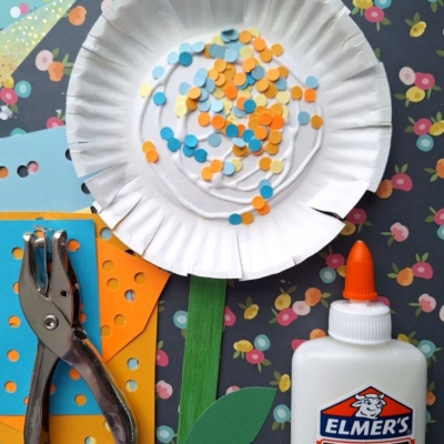A paper plate decorated as a flower with colorful paper confetti, surrounded by a hole punch, orange and yellow paper, green stem, glue bottle, on a flower-patterned background. @GLUEDTOMYCRAFTS is at the bottom.