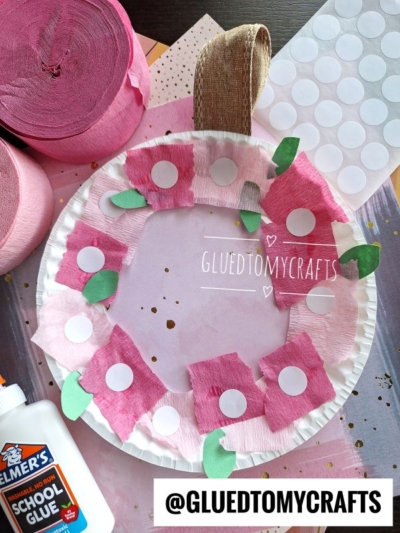 A paper plate apple craft decorated with pink and red crepe paper squares, green paper leaves, and white sticker dots—perfect for those searching for fun crepe paper craft ideas. Glue, pink crepe rolls, and a sticker sheet are nearby on the table.