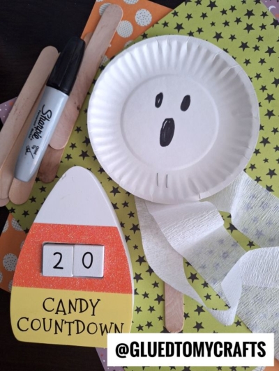 A paper plate ghost, craft supplies (Sharpie, craft sticks, toilet paper), Halloween-themed paper, and a candy corn countdown displaying “20” are arranged on a table—perfect for those seeking crepe paper craft ideas. Handle @GLUETOMYCRAFTS at the bottom.