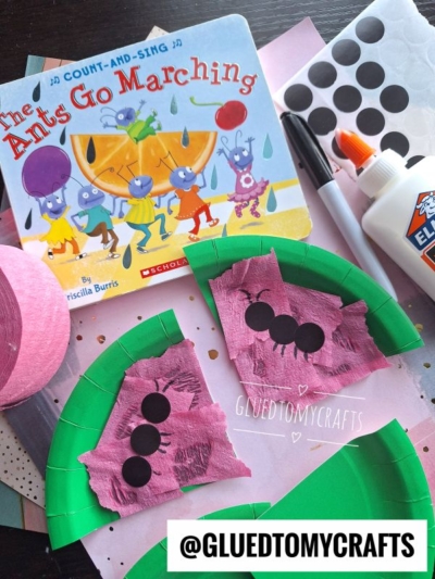 A children’s book titled The Ants Go Marching is surrounded by craft supplies, including pink and green paper pieces resembling watermelon slices for crepe paper craft ideas, black dot stickers, glue, scissors, and streamers. GLUEDTOMYCRAFTS watermark is visible.