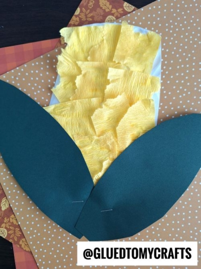 A corn-on-the-cob crepe paper craft idea made with torn yellow paper for kernels and two large green paper leaves, placed on autumn-themed paper. The handle @GLUEDTOMYCRAFTS is visible at the bottom.