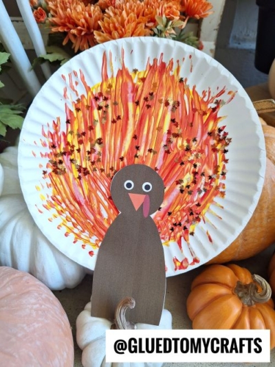A paper plate decorated with painted orange, yellow, and red streaks and star-shaped sequins to resemble a turkey’s tail, with a brown paper turkey body attached in front. White and orange pumpkins surround it.